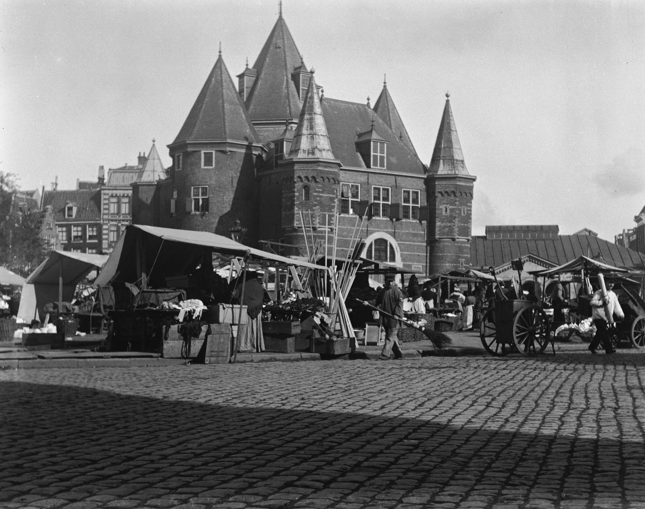 Market stalls, handcarts and a man sweeping the cobbles in front of the turreted Waag on the Nieuwmarkt in Amsterdam in 1902