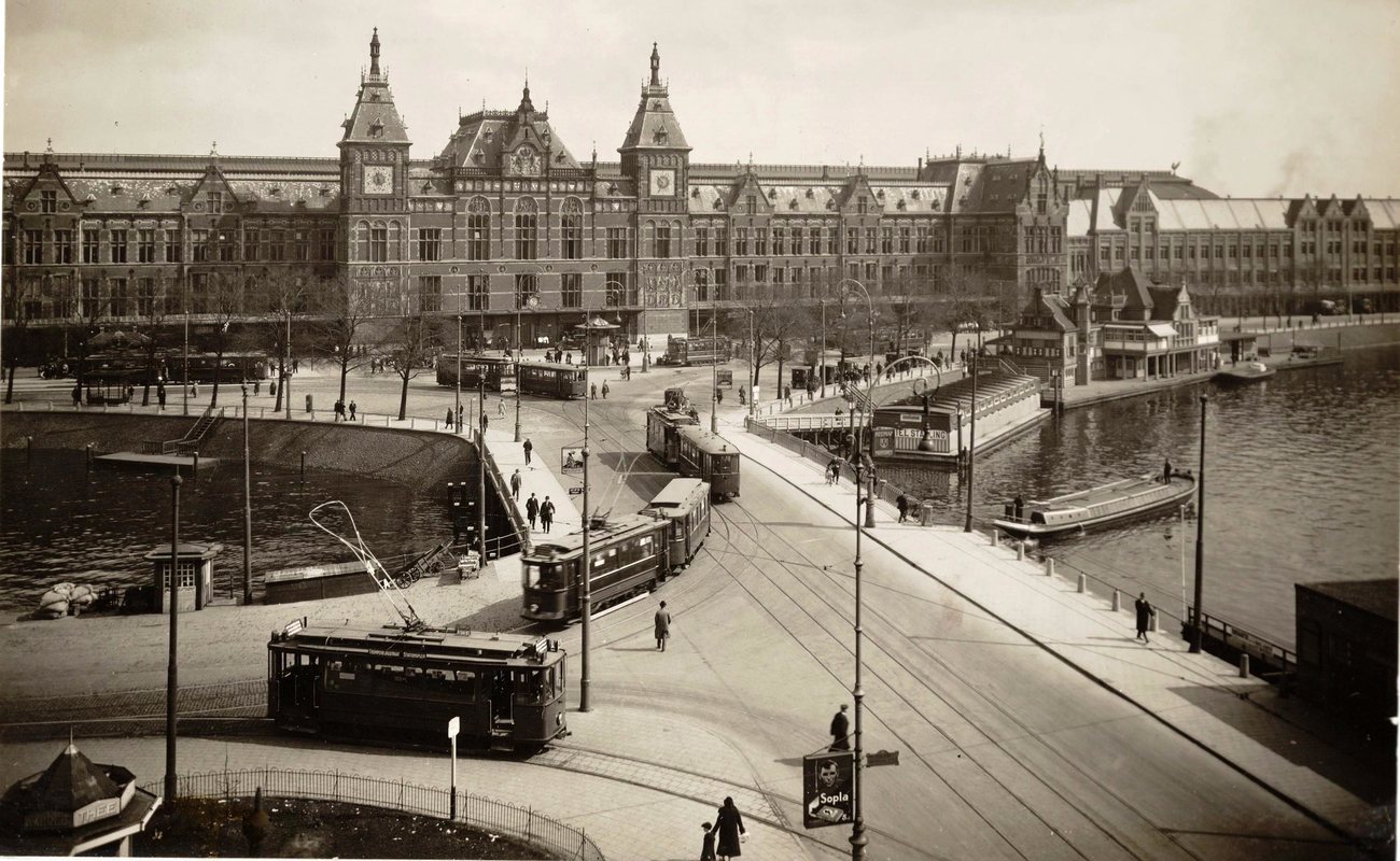 Trams curving across Stationsplein in front of Amsterdam's Centraal Station in the late 1920s, with boats moored on the water to the right