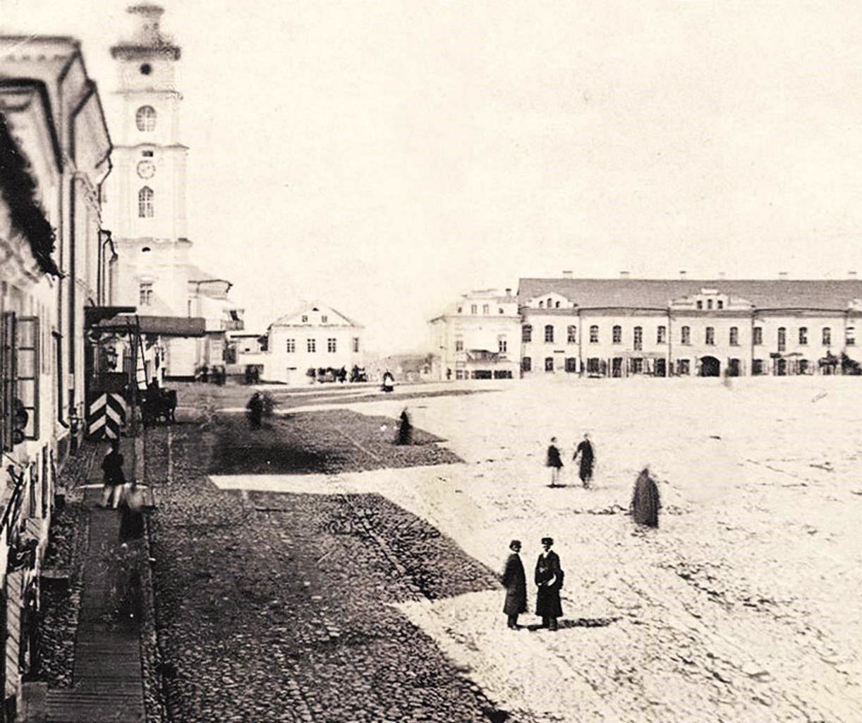 Men in long coats crossing the broad Cathedral Square in Minsk in 1865, a white clock tower on the left and a long two-storey arcade across the square