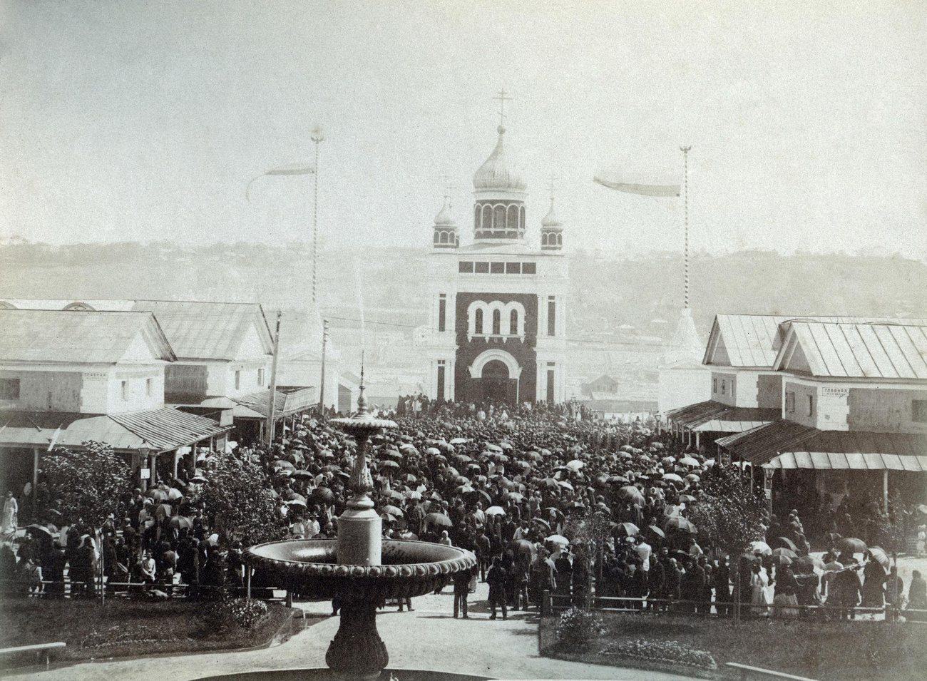 Crowds under umbrellas filling the fairground in front of the Makaryevskaya Chapel at the opening of the Nizhny Novgorod Fair in the early 1900s, a fountain in the foreground