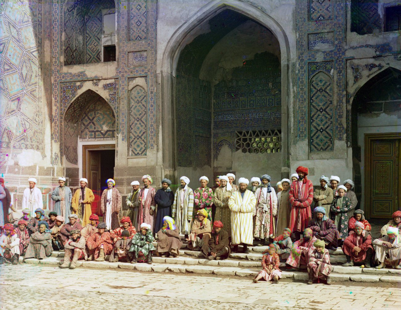 Men in striped and flowered robes and white turbans gathered on the steps of a tiled madrasa portal on the Registan in Samarkand, photographed in colour in 1911