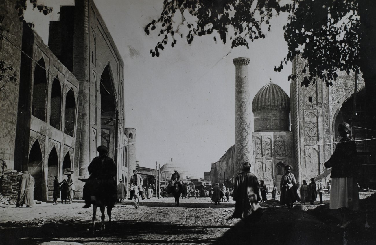 Donkey riders and people on foot on Yangi Rasta Street in Samarkand in the late 1920s, passing the tiled madrasas of the Registan, the Chorsu dome at the far end