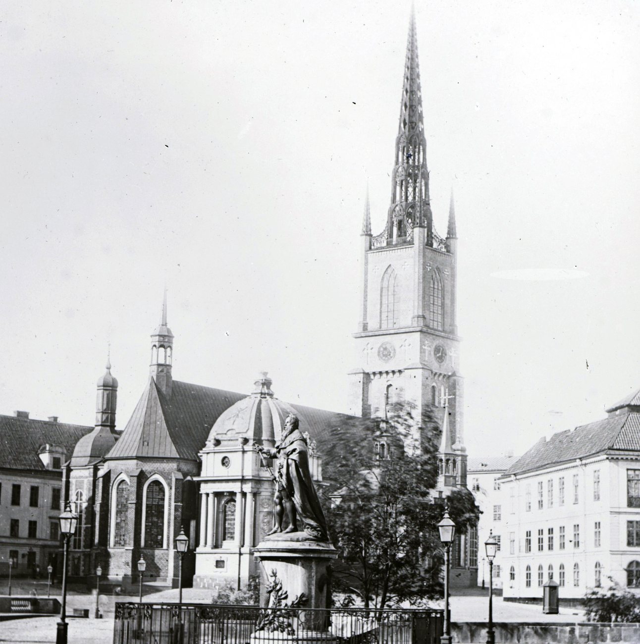 The openwork cast-iron spire of the Riddarholmen Church rising over a statue behind railings in Stockholm in 1862