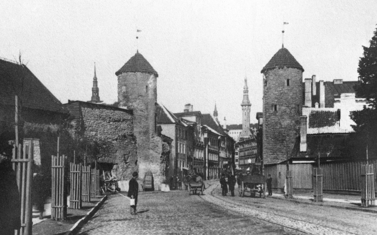 Carts and passers-by on the cobbled road between the two round towers of the Viru Gate in Tallinn around 1890, a spire rising beyond them