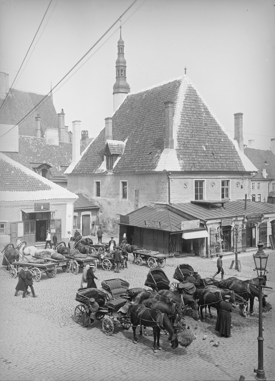 Horse cabs and loaded carts waiting on Town Hall Square in Tallinn in the early 1900s, a slender spire rising behind the rooftops