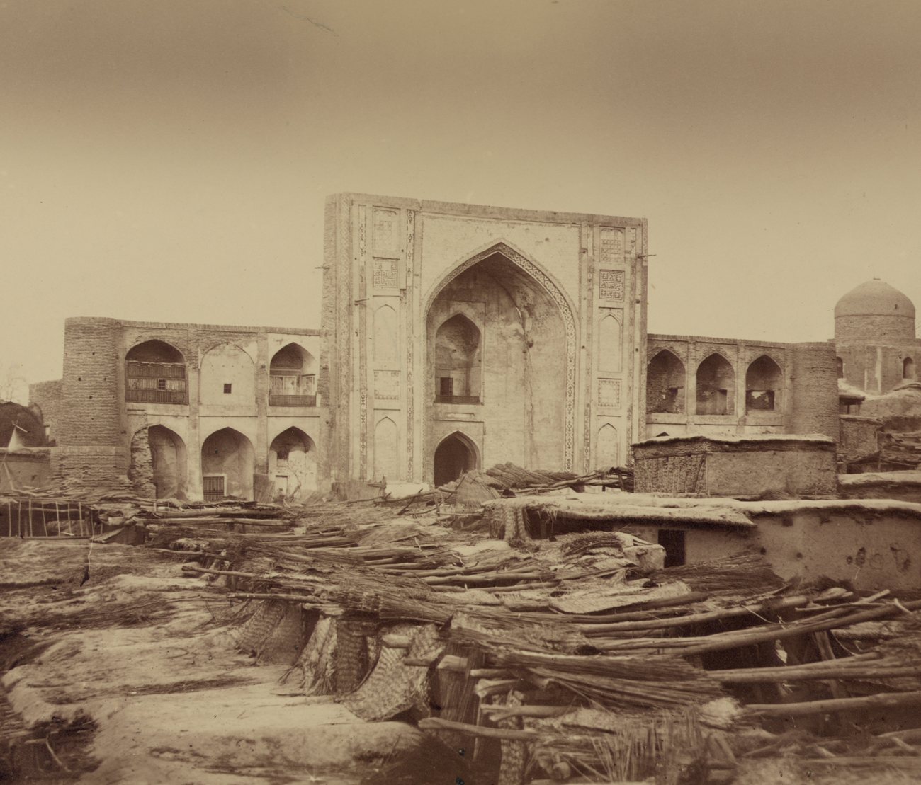 The tall portal and arcaded wings of the Kukeldash Madrasa in Tashkent in 1866, seen across reed-mat roofs and timber poles