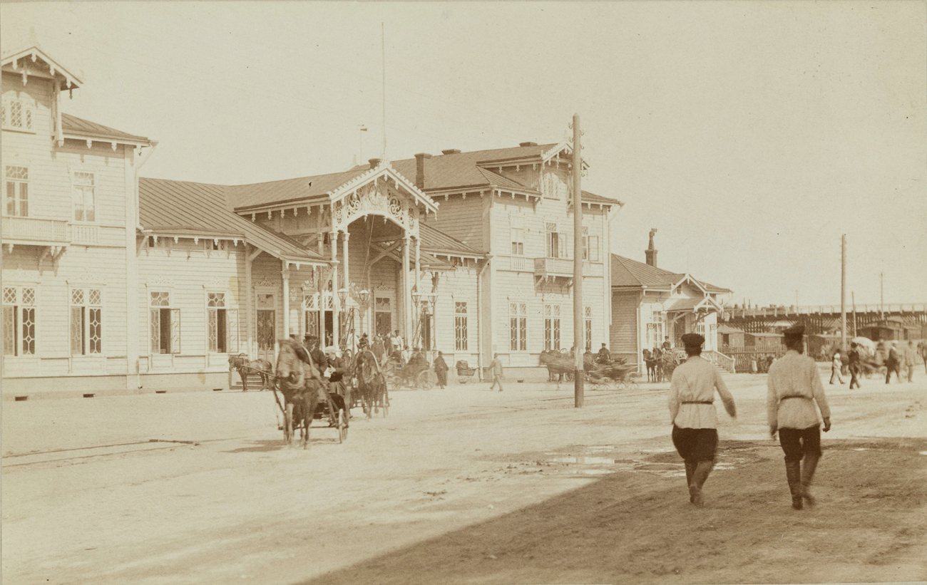 Horse cabs outside the wooden railway station in Vyborg in the early 1890s, two men in white belted tunics walking past